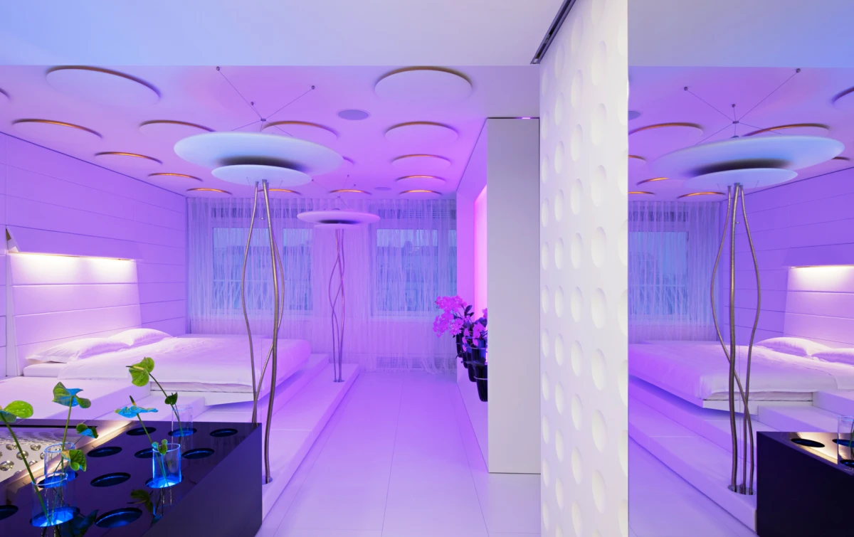 Futuristic spa suite with two white platform beds, purple ambient lighting, circular ceiling panels, and glass vases of plants.