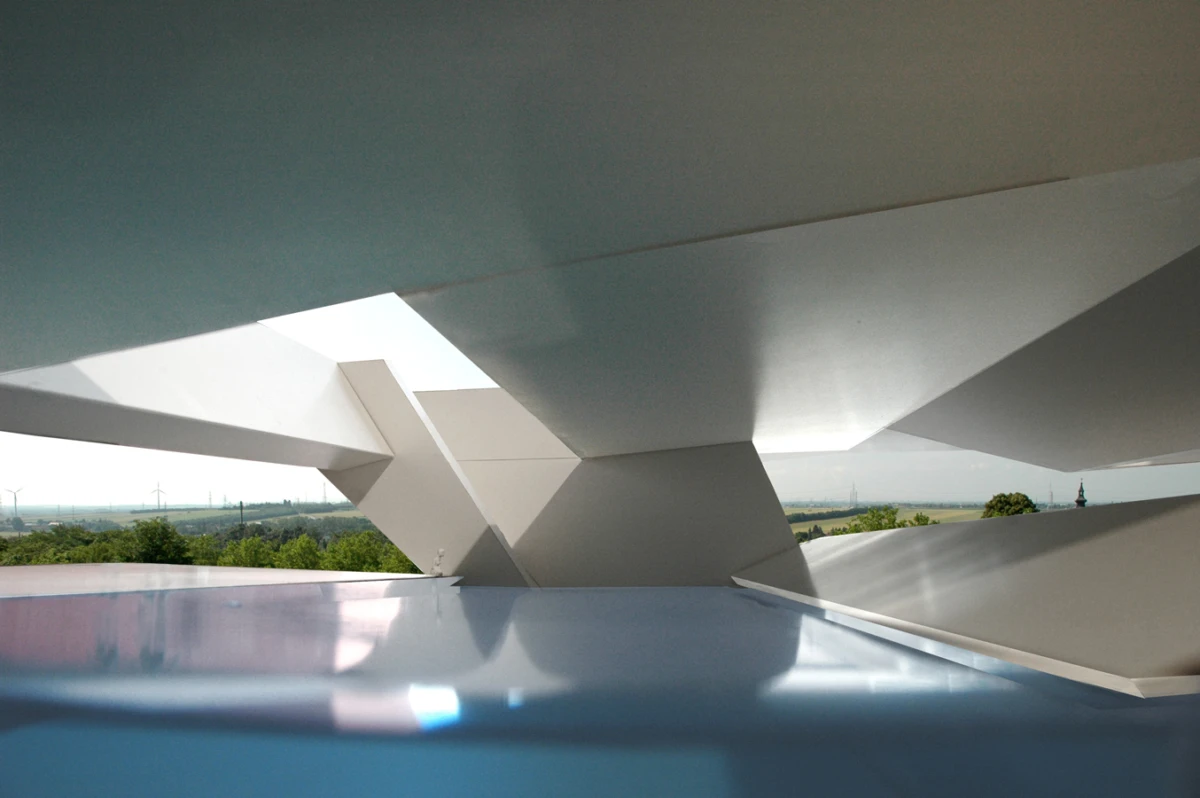 Modern white architectural structure with angular planes, viewed from below, overlooking a green countryside with wind turbines.