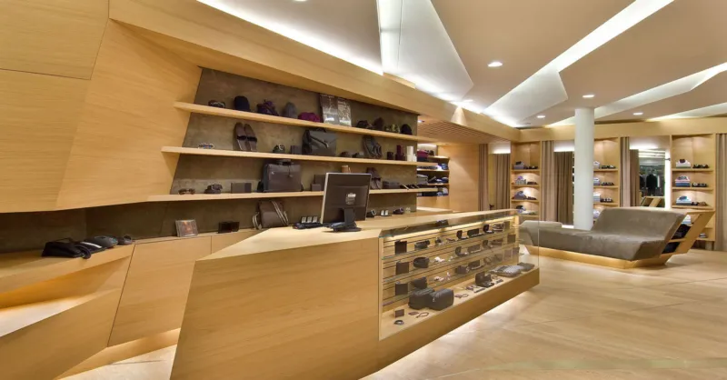 Modern wooden boutique interior with a glass eyewear display counter; wall shelves hold sunglasses and handbags, seating area to the right.