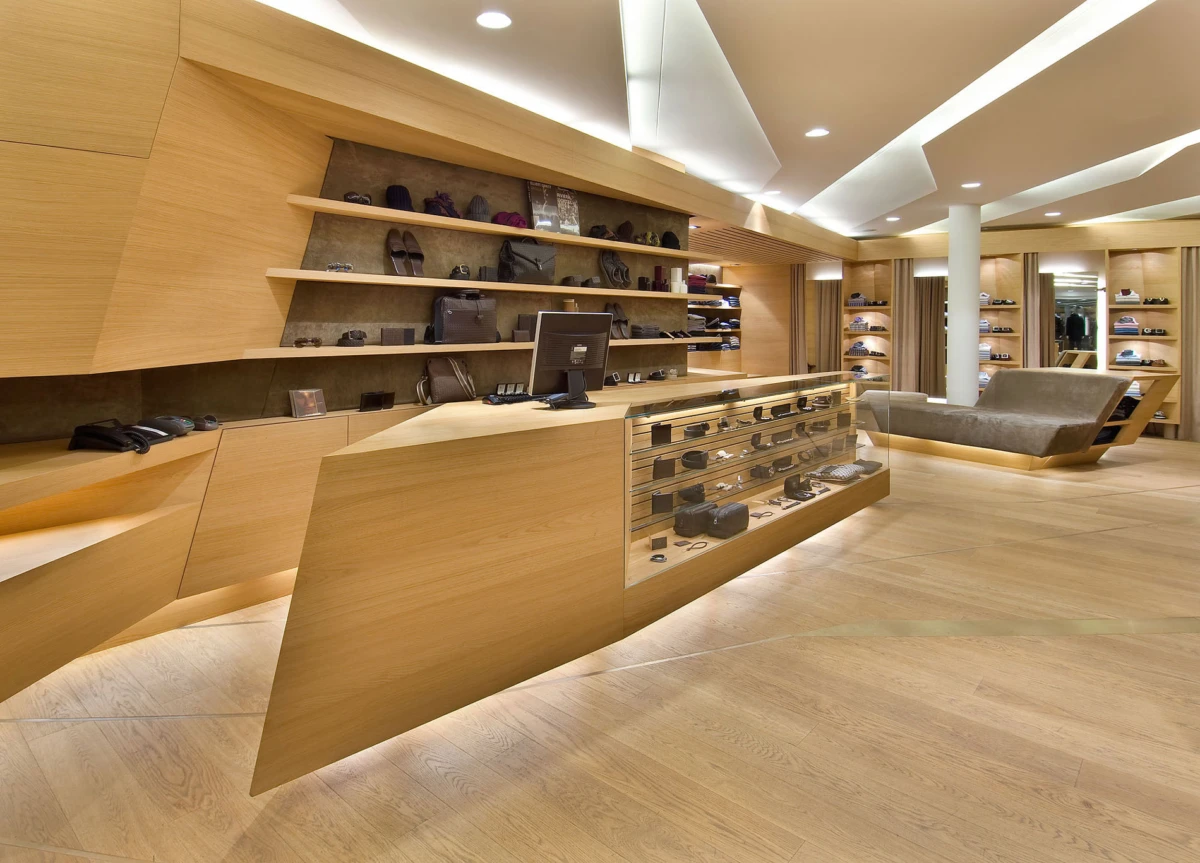 Modern wooden boutique interior with shelves of handbags and wallets, a glass display counter, and a gray seating area.