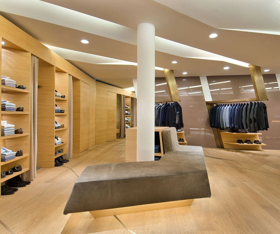Modern men's clothing store interior with suit jackets on racks, folded shirts on wooden shelves, and a gray upholstered bench center.