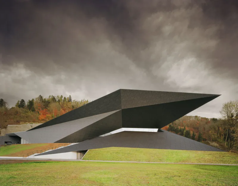 Contemporary angular building with layered wedge-shaped roofs on a grassy hill under a cloudy sky.