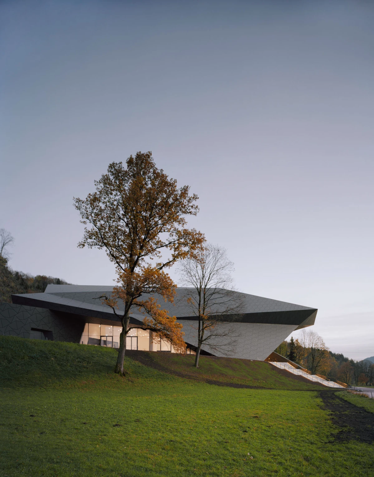 Angular modern building on a grassy hill, two autumn trees in foreground, twilight sky.