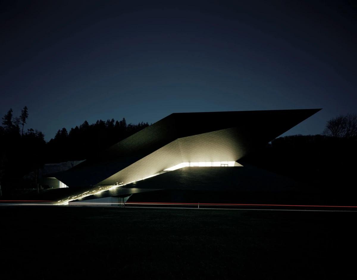 Night view of a modern architectural structure with a large slanted roof and illuminated ramp, with a red taillight streak across the foreground
