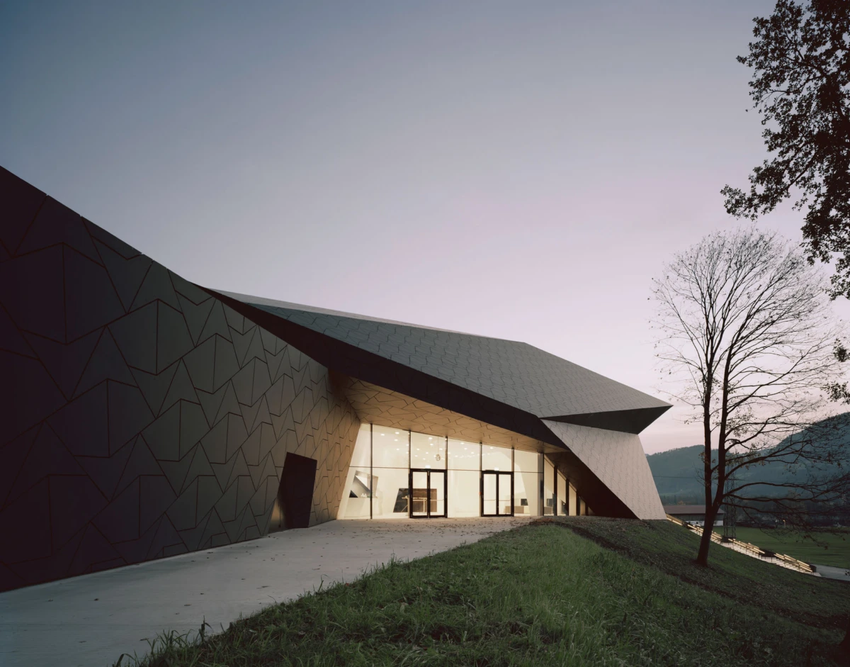 Modern angular building with dark geometric exterior and glass entrance, standing on a grassy slope beside a leafless tree at dusk.