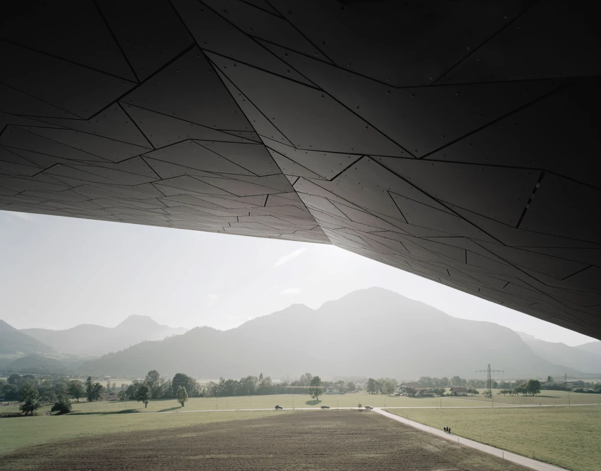 Polygonal metal canopy roof dominates the foreground; rural landscape with fields, a road, trees, and hazy distant mountains.