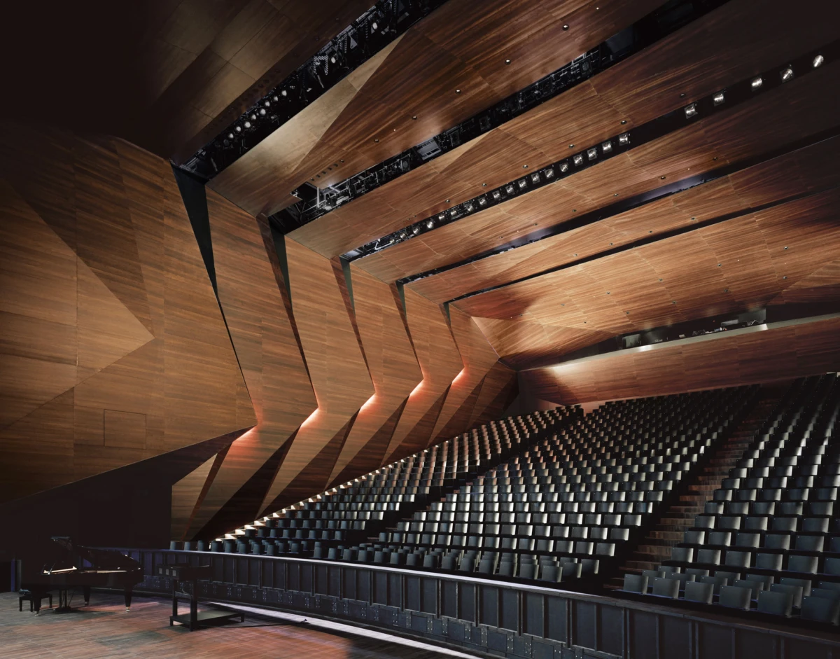 Interior of an empty modern concert hall with angular wooden walls and tiered seating; a grand piano on stage.