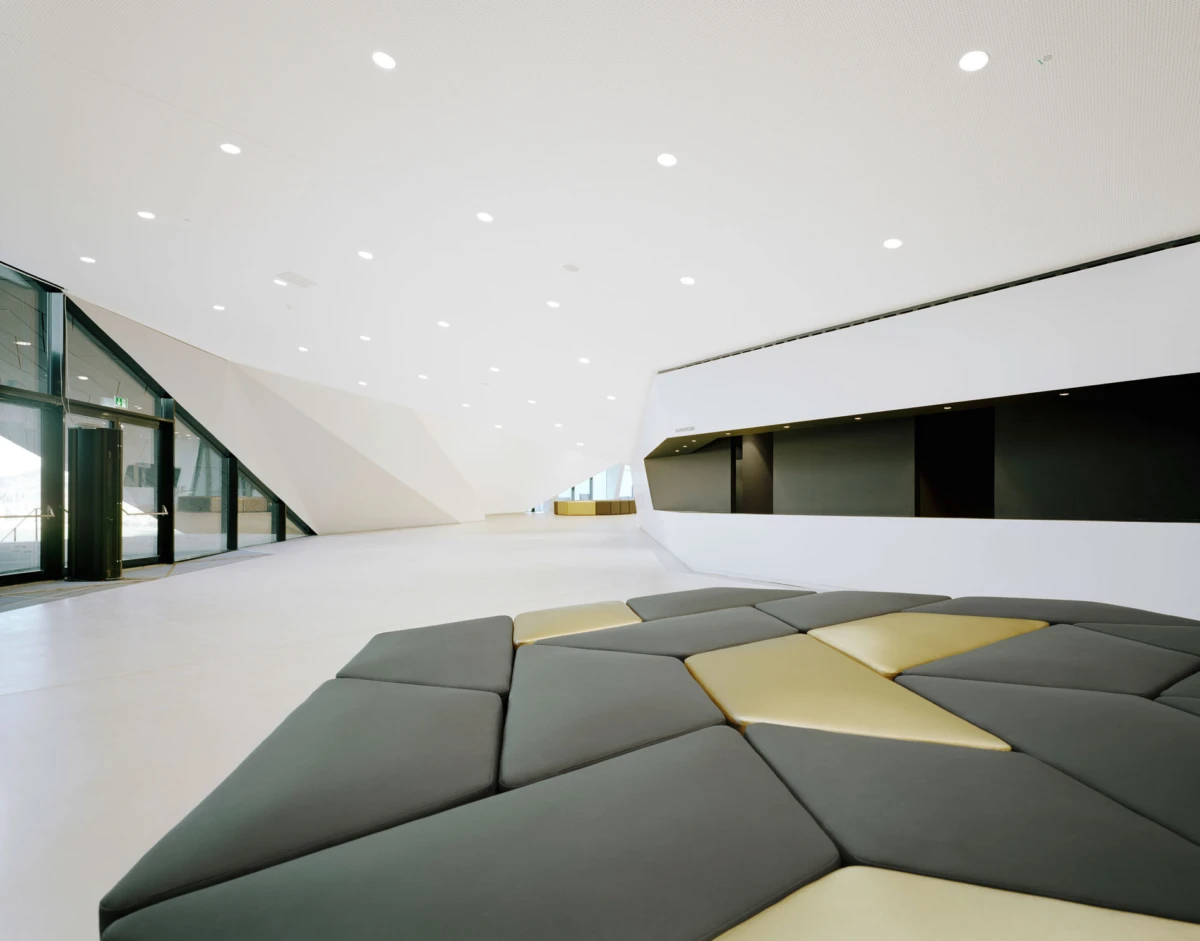 Empty futuristic lobby with white curved walls, bright recessed lights, and gray and yellow modular cushions arranged in a circular pattern.