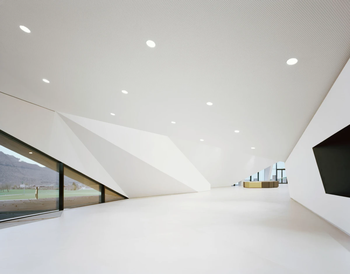 Minimalist white interior with angular walls and a large glass wall; empty lounge overlooking fields and mountains.