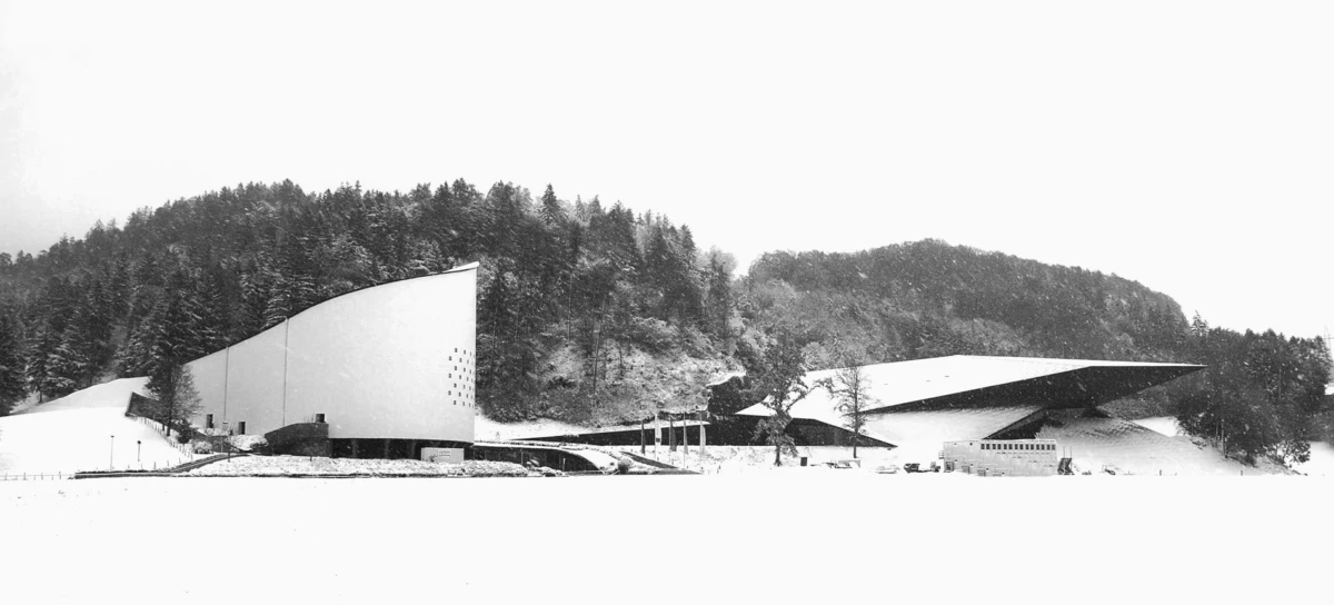 Two modernist pavilions with angular roofs in a snowy field, backed by a dense pine forest.