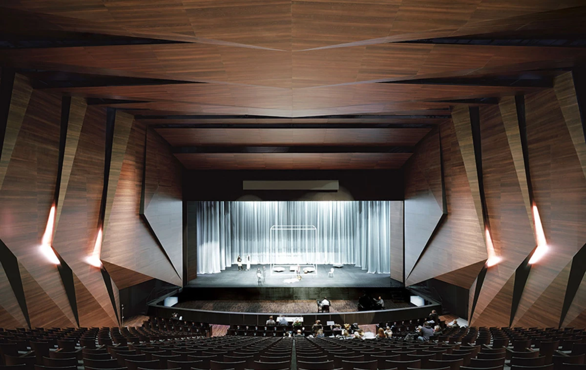 Interior of a modern wood-paneled theater auditorium; performers rehearse on a stage with white curtains.