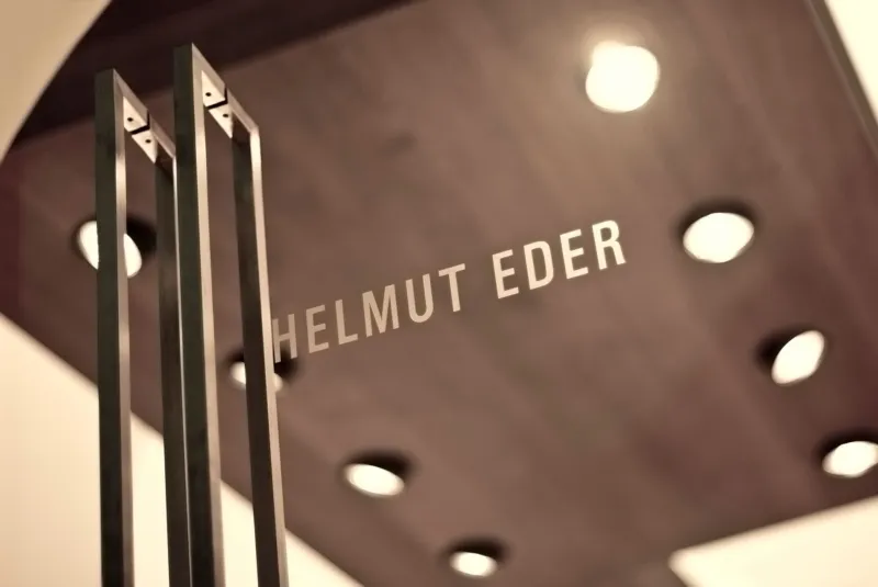 Three slender metal bars in foreground; a blurred wooden panel with the text HELMUT EDER and circular lights in the background.