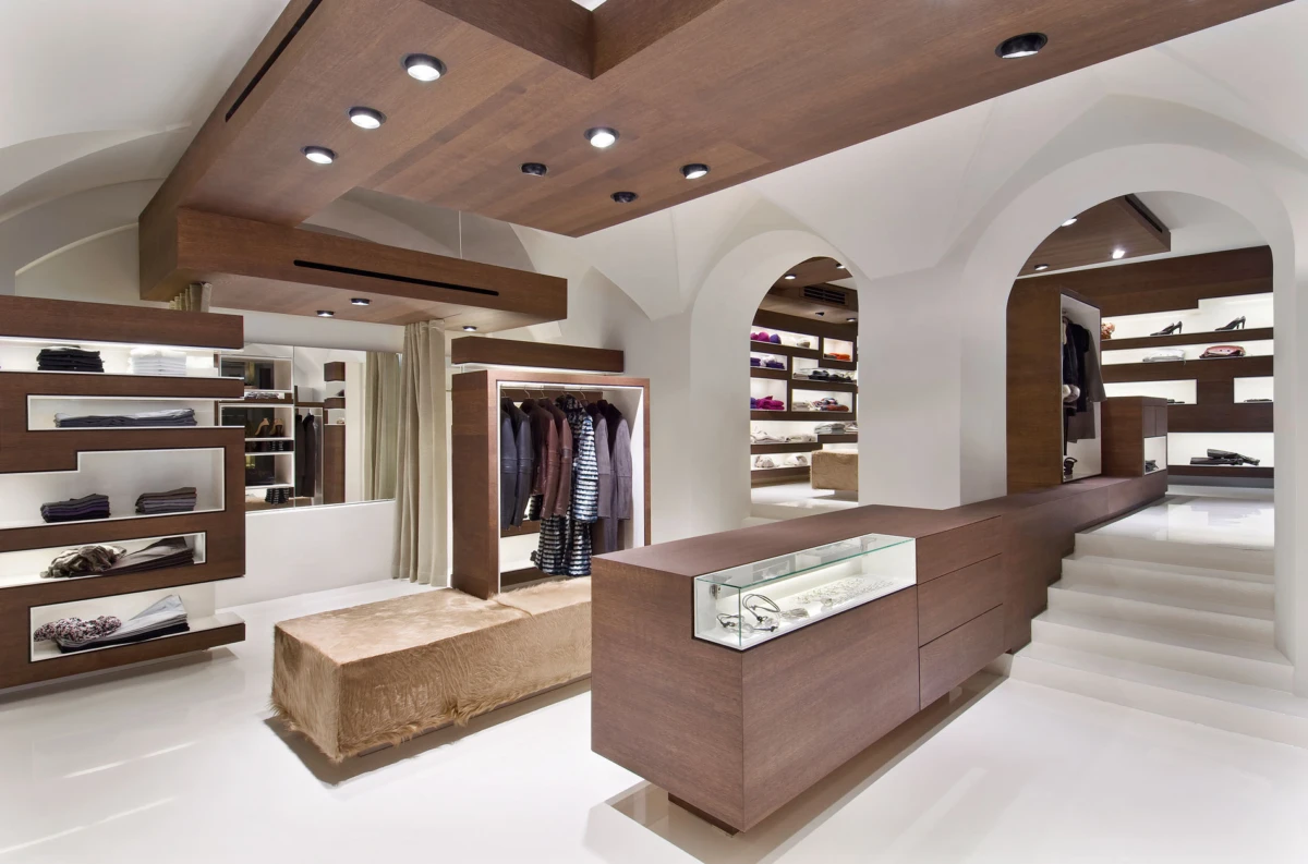 Interior of a modern clothing boutique with a long wooden sales counter, glass display case, arched white walls, and clothing on racks.