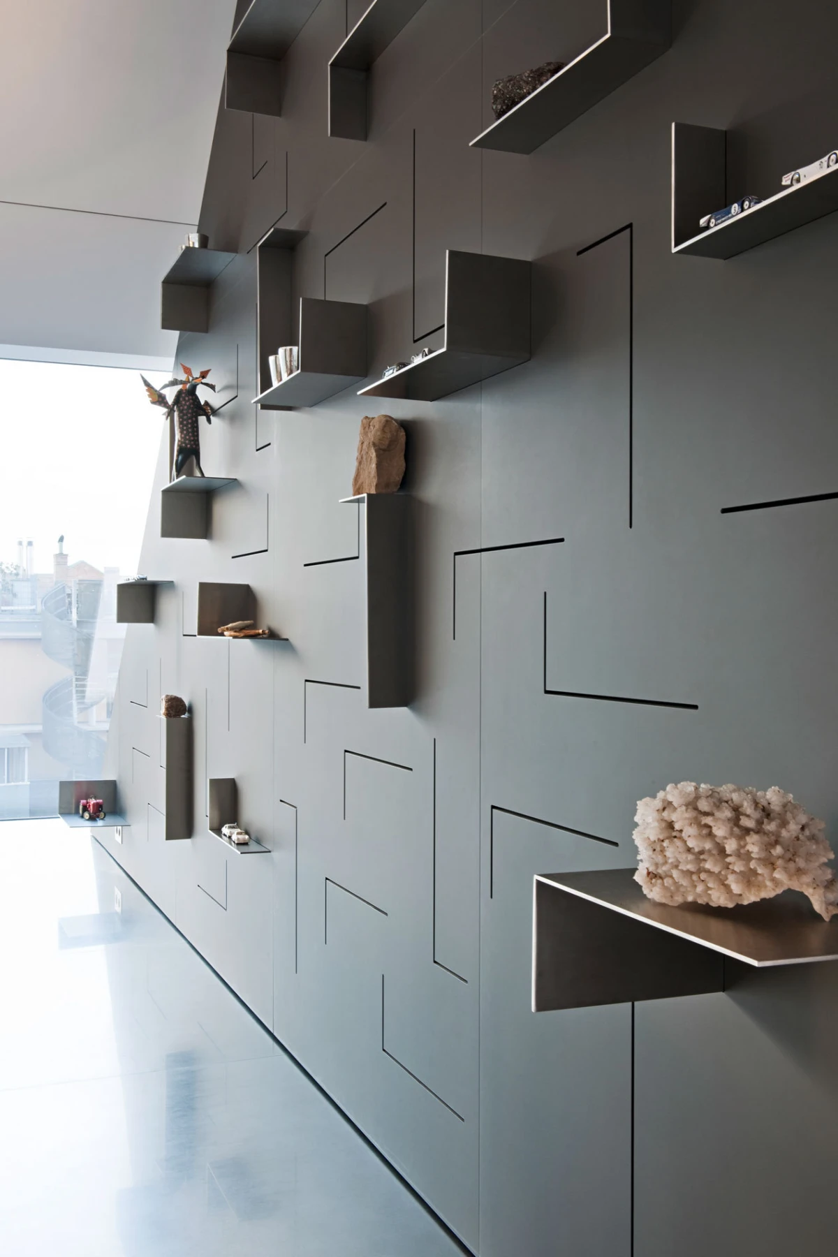 Gray wall with geometric, protruding shelves displaying small objects; a sculptural figure and coral on display in a modern gallery interior.