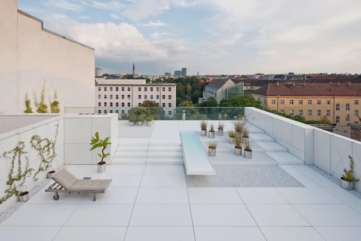 Unoccupied beige cushioned lounge chair on a white rooftop terrace with planters and steps, overlooking an urban skyline.