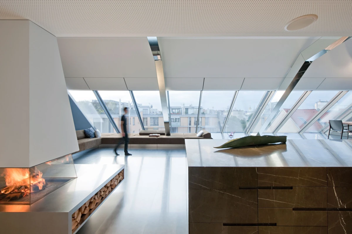Person walking through a modern open living area with a fireplace, bench seating, and a marble kitchen island under angled skylight windows.