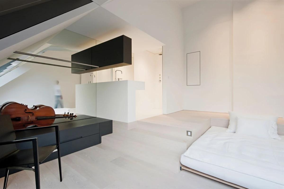 Modern loft apartment with a sloped ceiling; violin rests on a black cabinet beside a desk chair, white bed nearby.