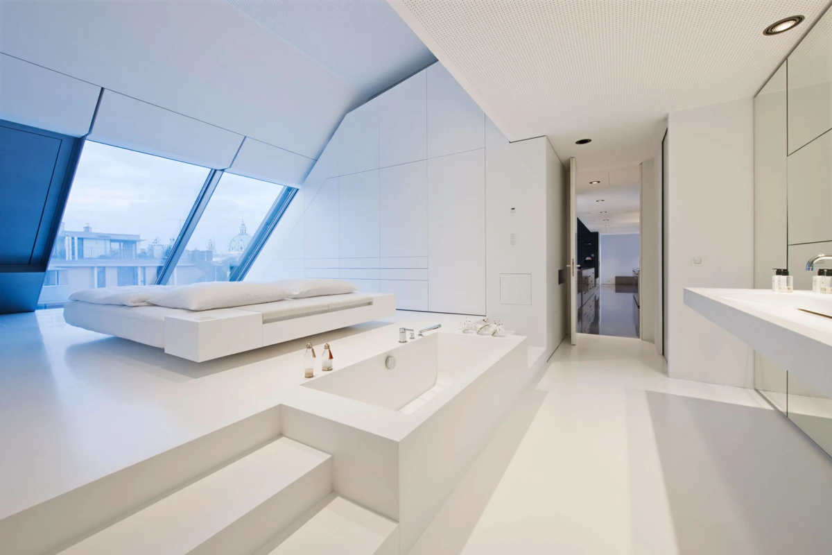 White minimalist bedroom with an elevated platform bed beside slanted glass windows; built-in bathtub with steps and a long vanity.