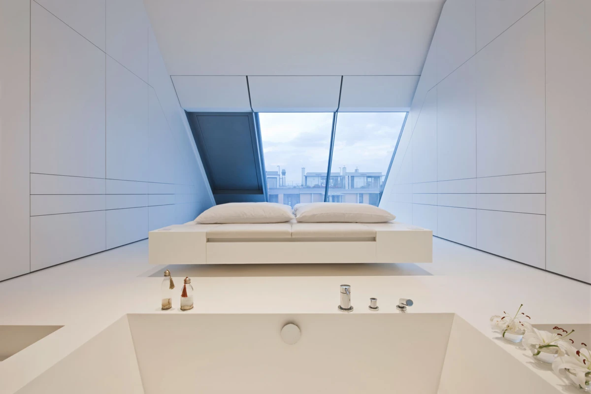 Minimal white bedroom with a low platform bed under a large angled skylight; foreground shows a bathtub with taps and white flowers.