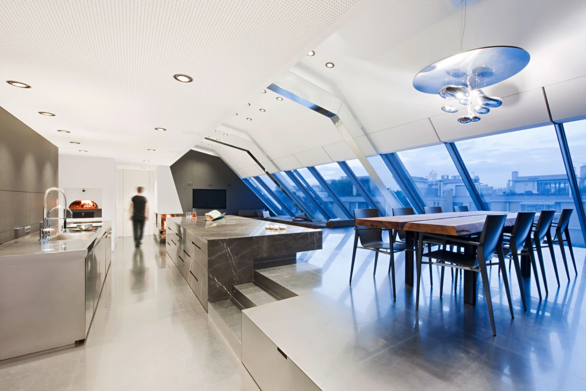 Modern open-concept kitchen with marble island and stainless appliances; dining table beside slanted glass walls; a person walks in the background.