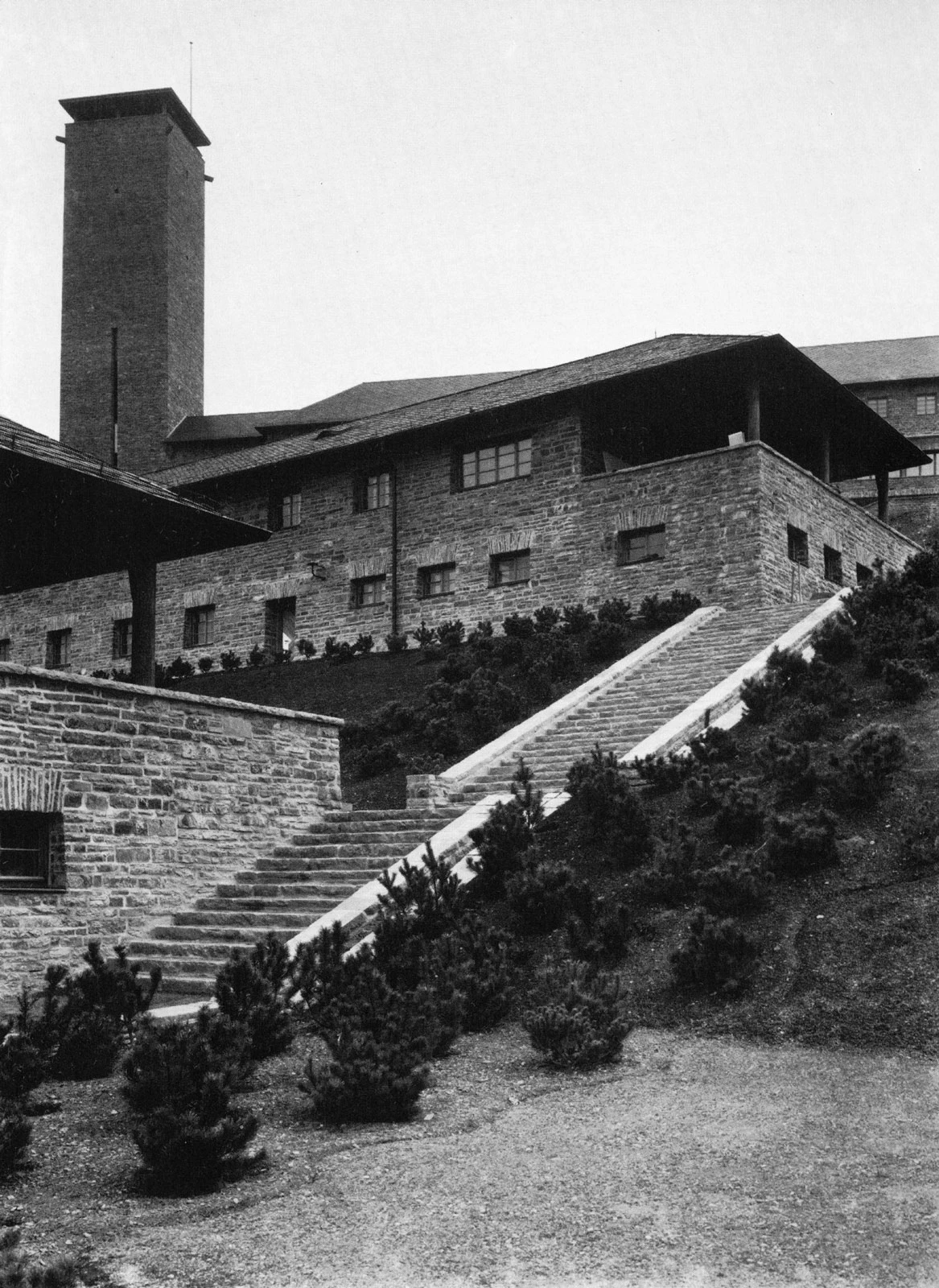Stone hillside building with a tall brick tower and a long staircase ascending the slope; shrubs in the foreground.