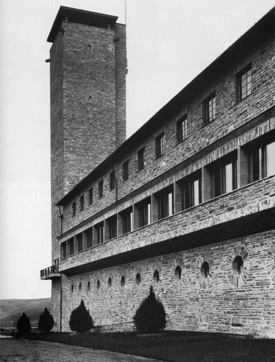 Stone fortress with a tall square tower on the left and a long wall of rectangular windows; circular openings along the base.