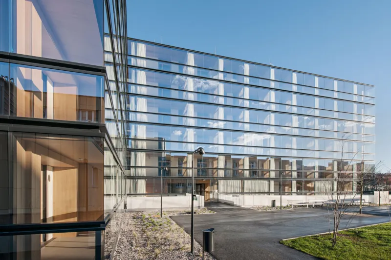 Glass-clad office building with reflective facade, empty driveway, and landscaped foreground on a sunny day.