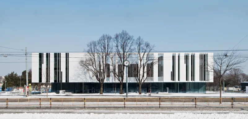 White modern office building with vertical black window panels and a central clock, flanked by leafless trees on a snowy street.