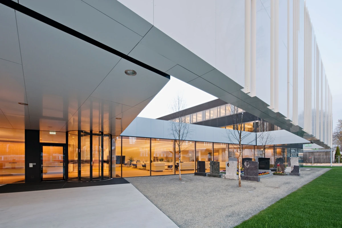 Modern office building exterior with glass walls and an overhanging canopy; memorial stones and trees in a gravel forecourt.
