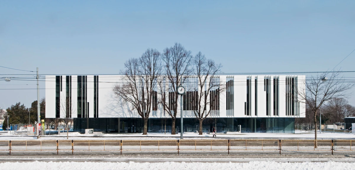 White modern office building with vertical black window panels and a central clock, flanked by leafless trees on a snowy street.