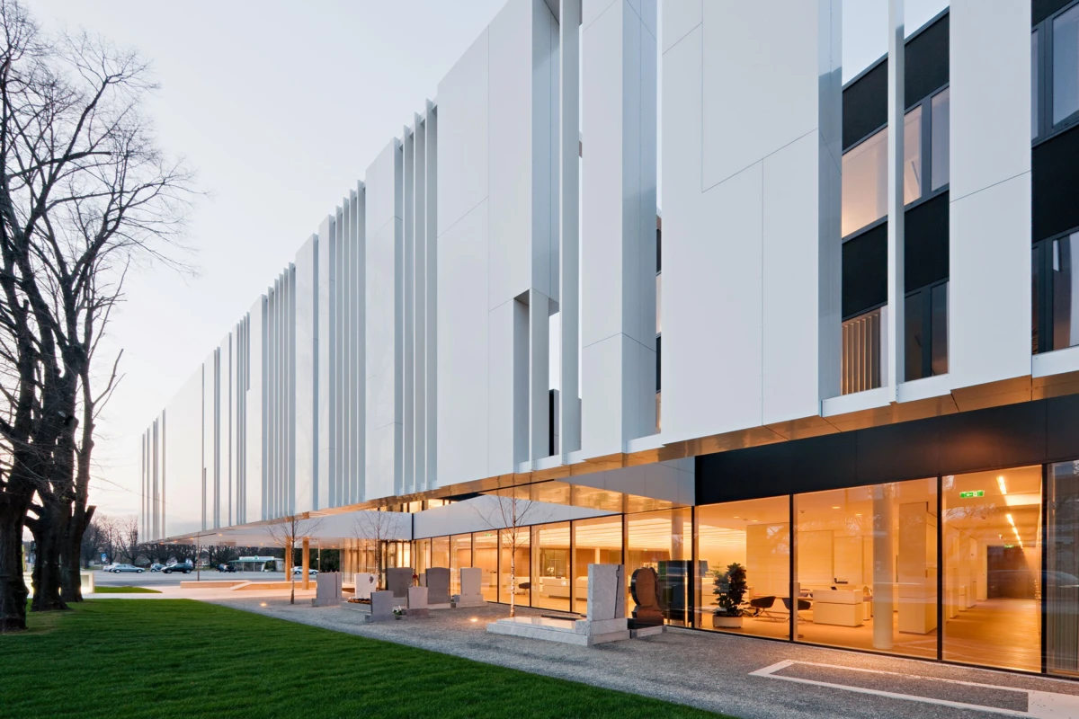Exterior of a modern white office building with vertical fins, glass storefronts, outdoor seating, on a grassy lawn with leafless trees at dusk.