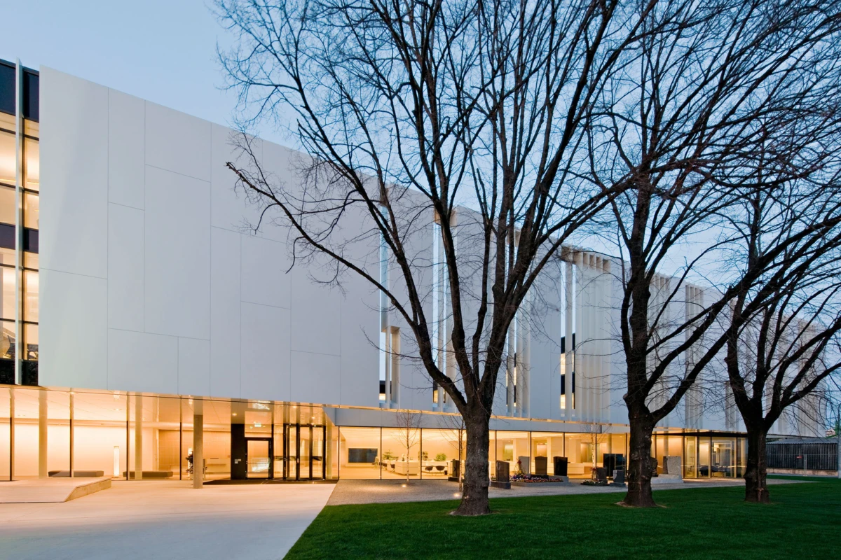 Modern white-facade building with glass entrance at dusk, leafless trees in foreground and green lawn.