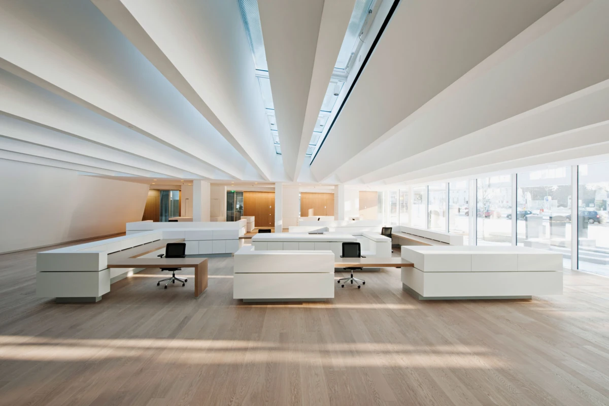 Unoccupied open-plan office with white modular desks, laptops, and glass walls; bright, airy space with wooden floor and skylight beams.