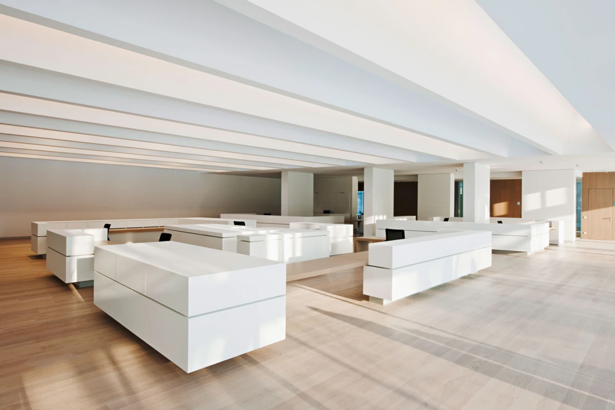 Modern white office reception area with low rectangular desks and black chairs in a sunlit, open minimalist space.