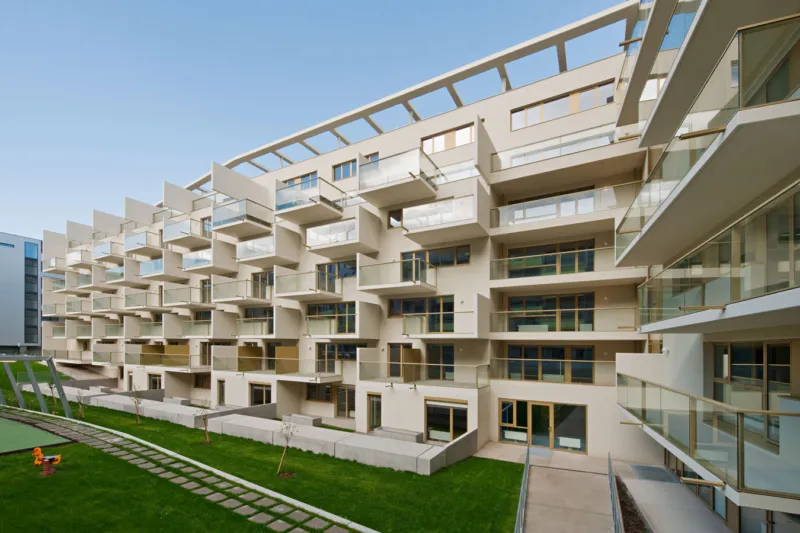 Modern beige apartment complex with multiple glass balconies around a green courtyard; paved path and a small playground in the foreground.