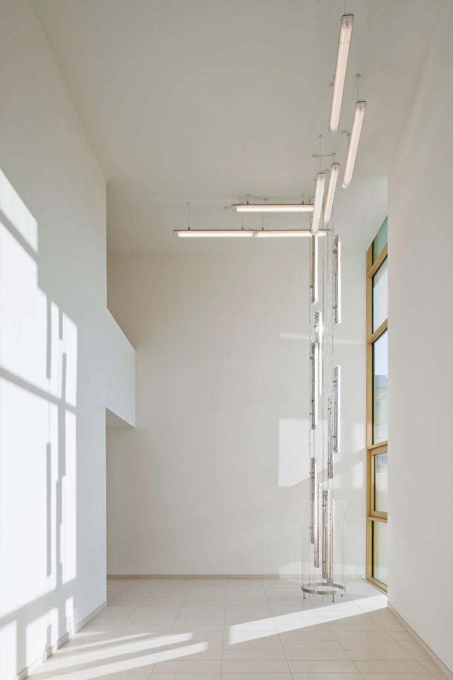 Bright white gallery with a tall suspended glass sculpture, rectangular prisms along a central pole, near a large window.