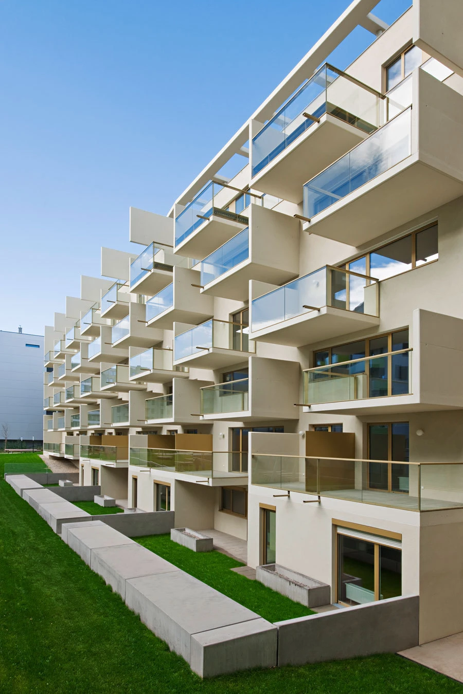 Modern beige apartment building with glass balconies, multi-story, overlooking a grassy courtyard with concrete walkways.