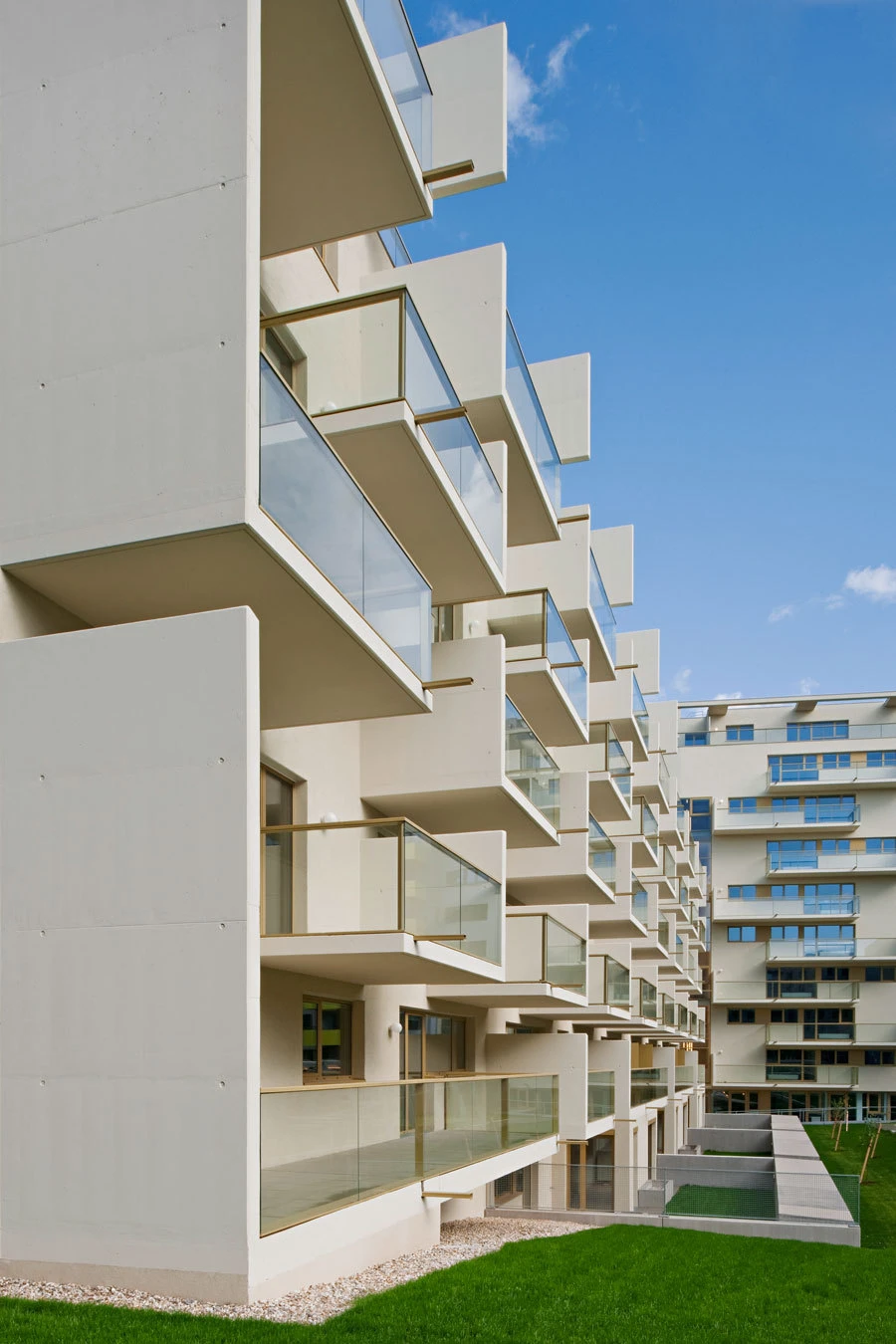 Modern white concrete apartment building with stacked glass balconies along a green lawn under a blue sky.