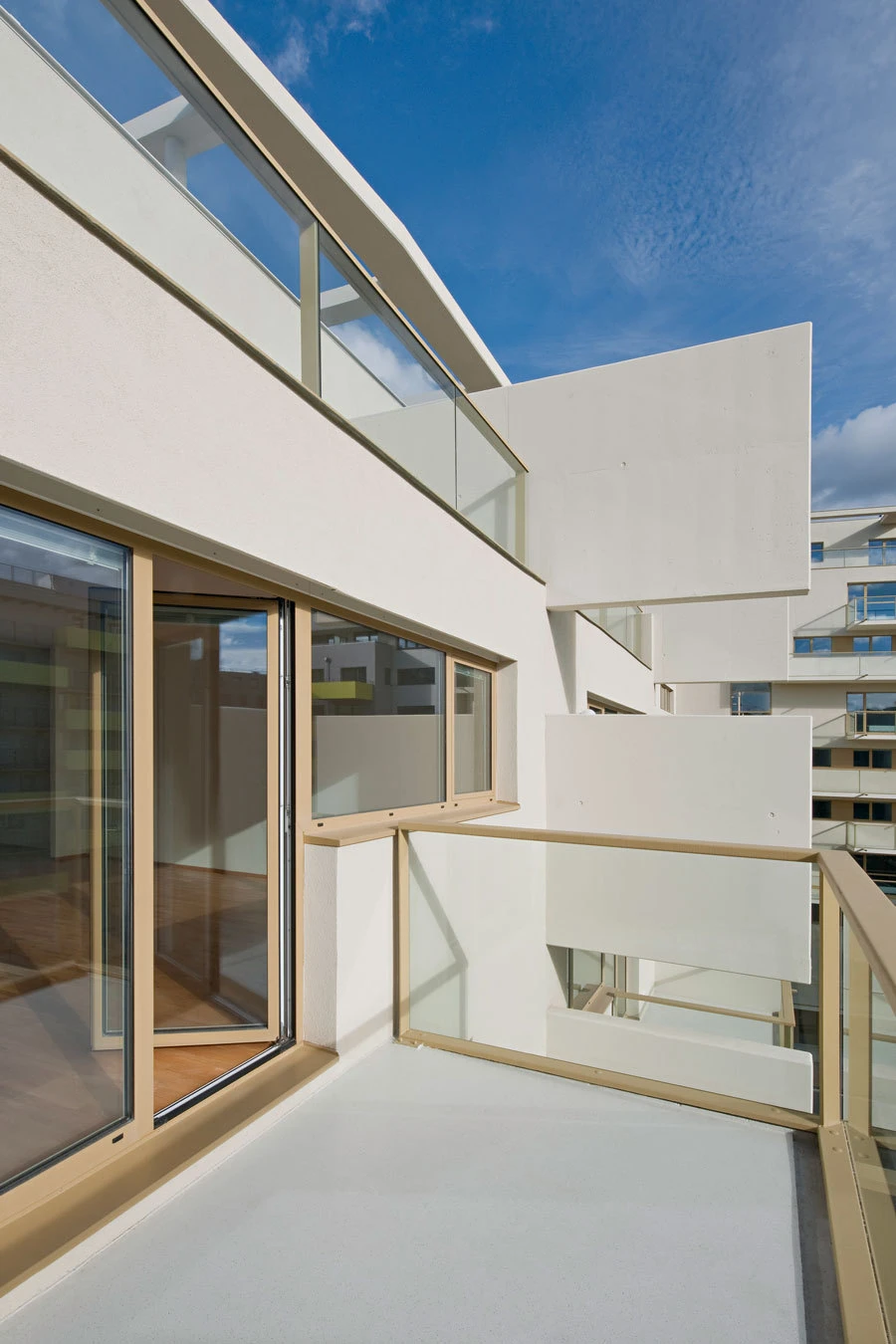 Empty balcony of a modern white apartment building with glass railing and sliding doors, under a bright blue sky.