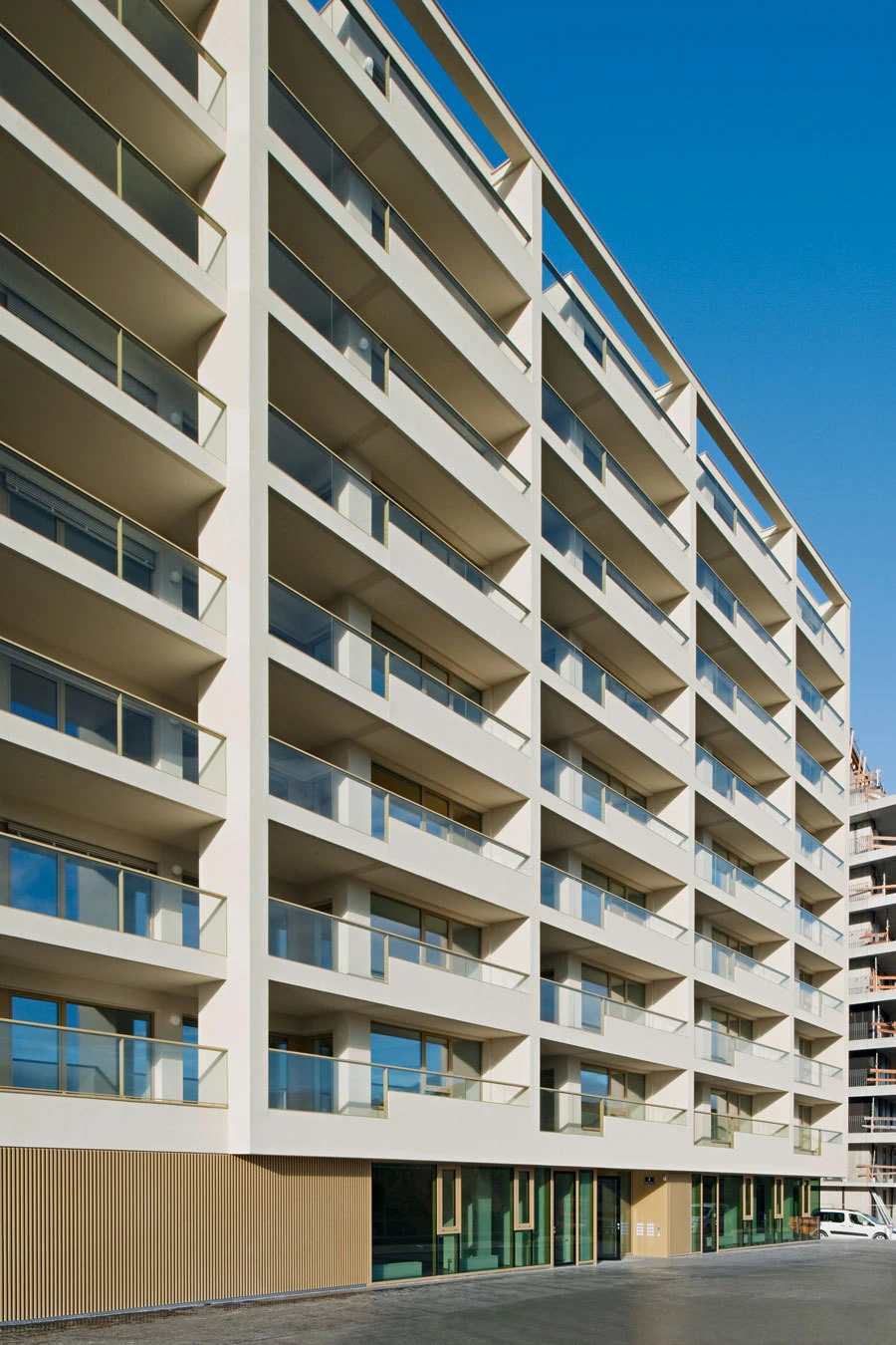 Modern white apartment building with uniform balconies along a street; glass storefronts on the ground floor under a blue sky.