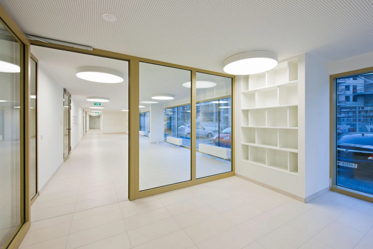 Empty office corridor with glass partitions, white tile floor, circular ceiling lights, and built-in shelving by a large window.