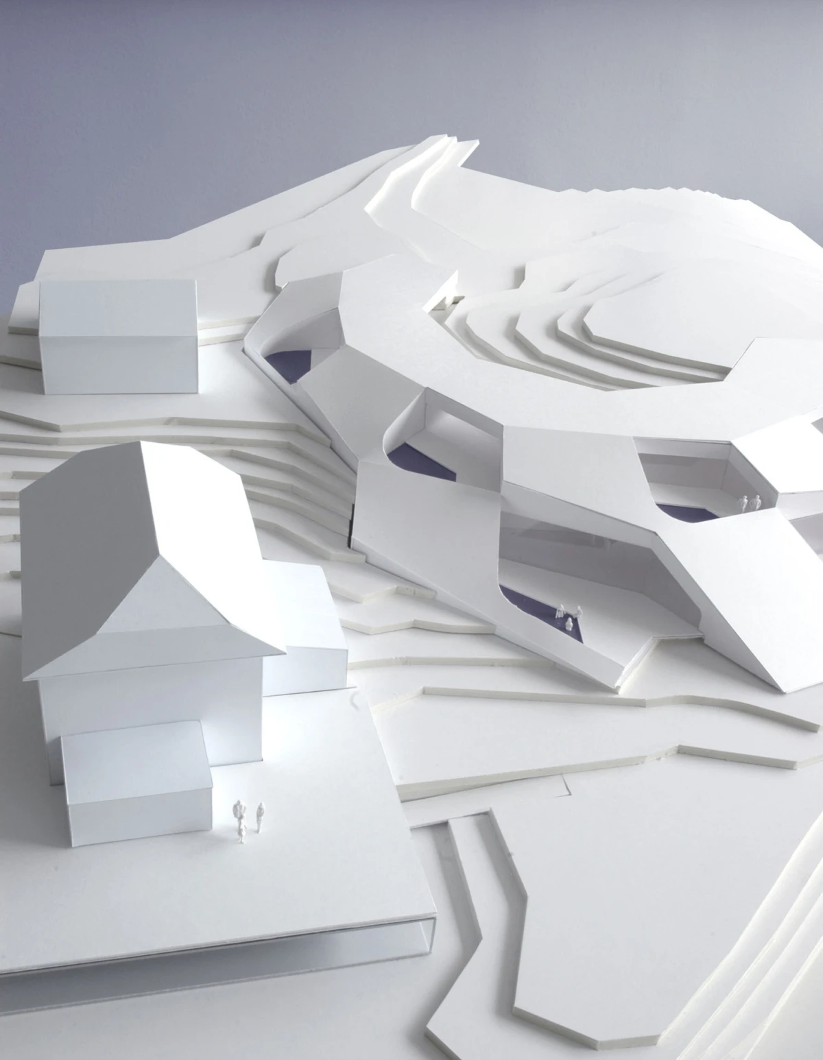 White paper architectural model of a terraced landscape with a small house and two tiny figures.