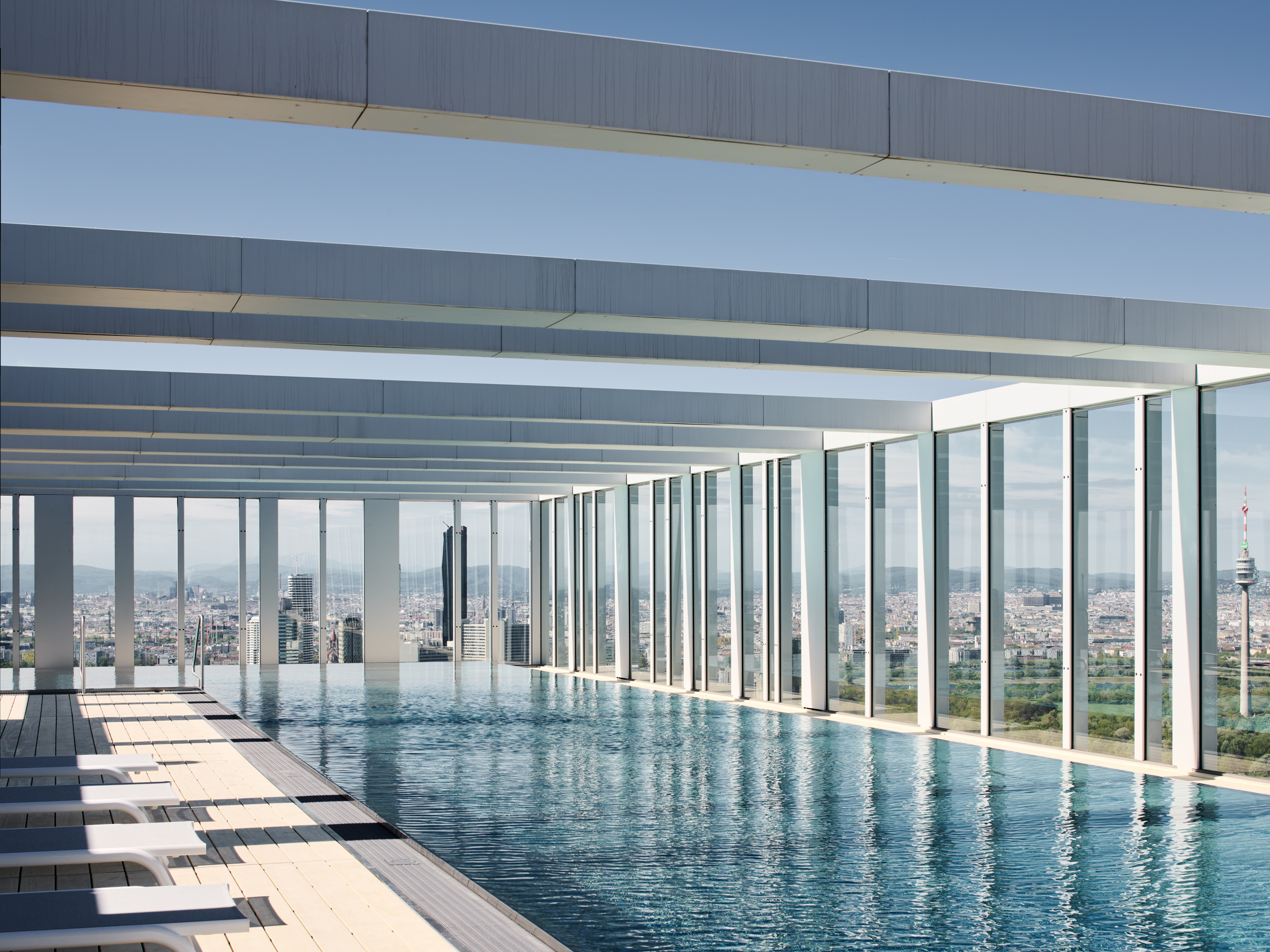 Rooftop infinity pool with glass walls overlooking a city skyline; white lounge chairs line the pool deck.