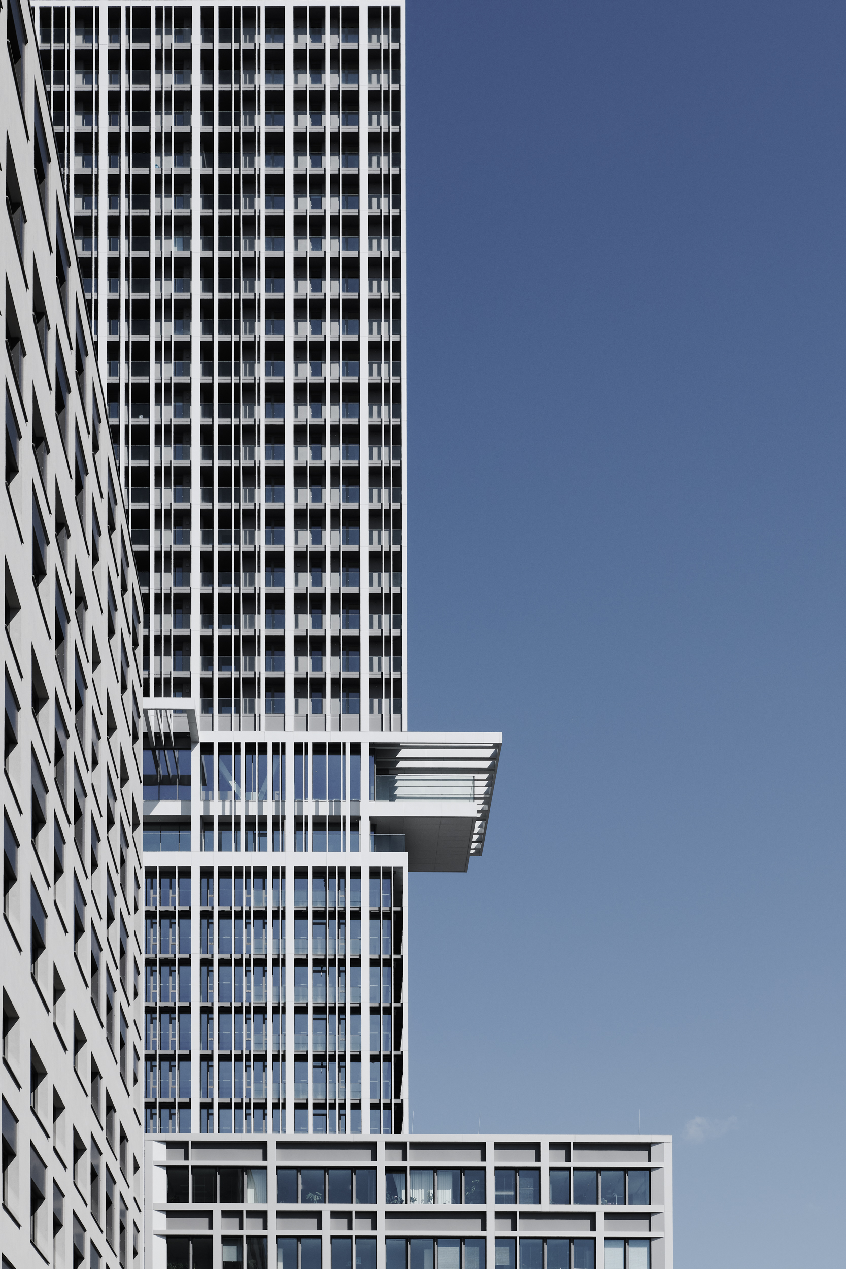 Tall modern glass office tower with a grid of windows and a cantilevered platform extending from its midsection against a clear blue sky.