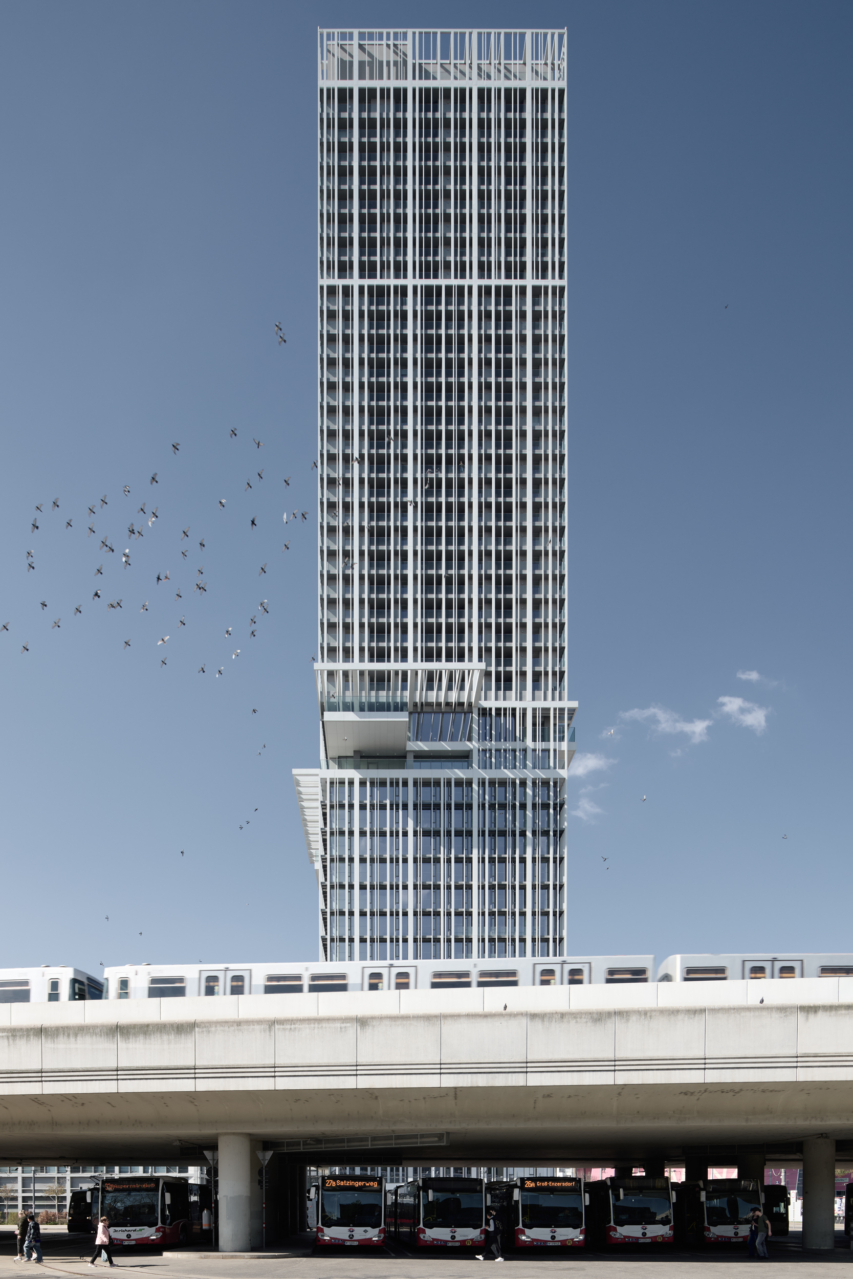 Glass-clad skyscraper with a stepped lower section; flock of birds in the sky; elevated transit deck with buses beneath in a city setting.