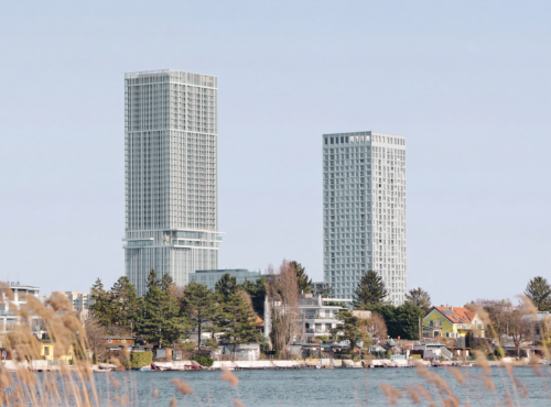 Two tall modern high-rise buildings rise above a residential waterfront with trees, reeds in the foreground, and houses along the shore.