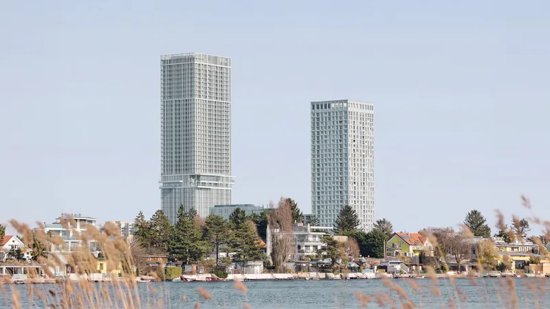 Two tall modern high-rise buildings rise above a residential waterfront with trees, reeds in the foreground, and houses along the shore.