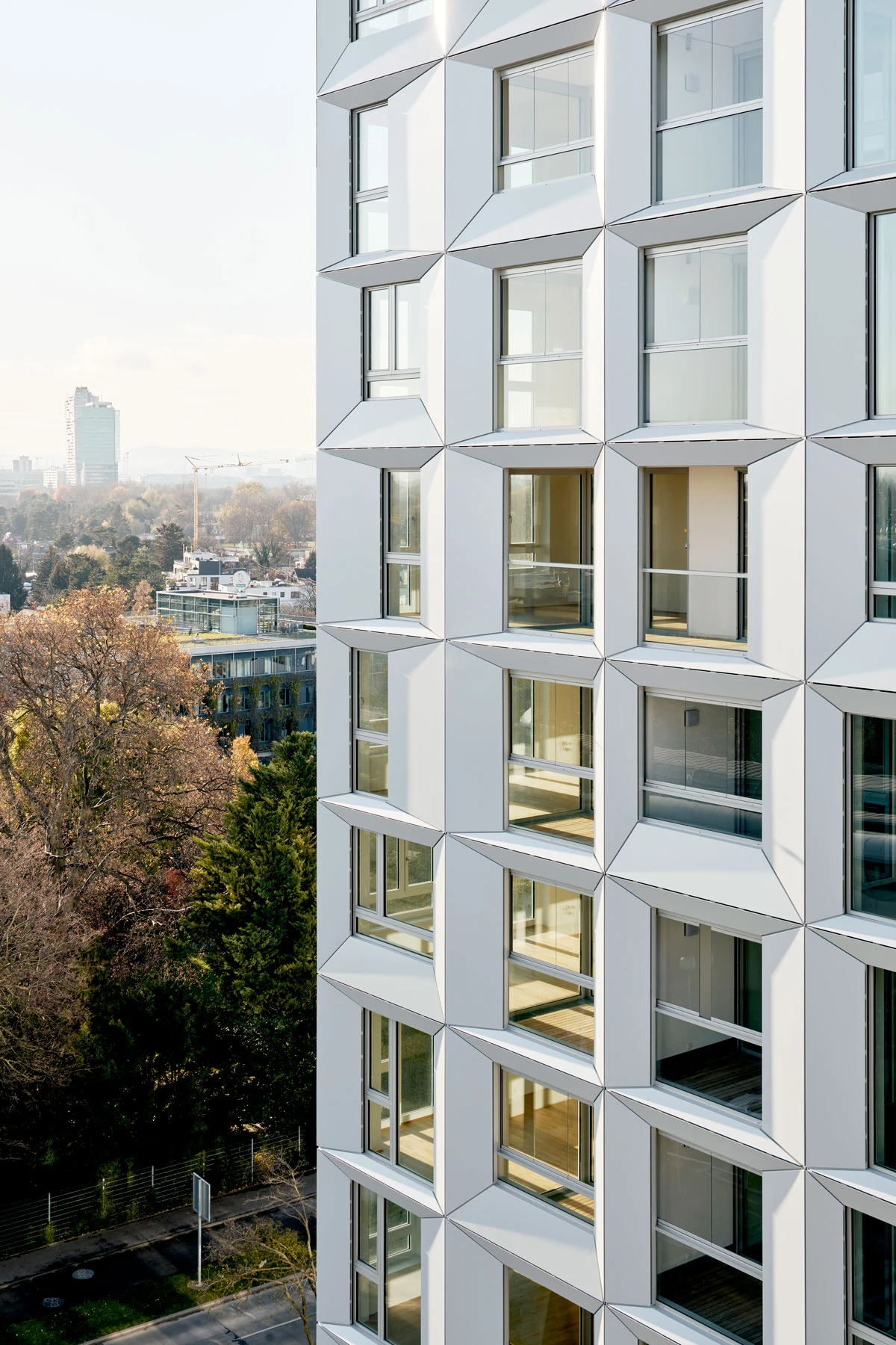 White high-rise with a diamond-patterned facade and large square windows; sunlit cityscape and trees in the foreground.