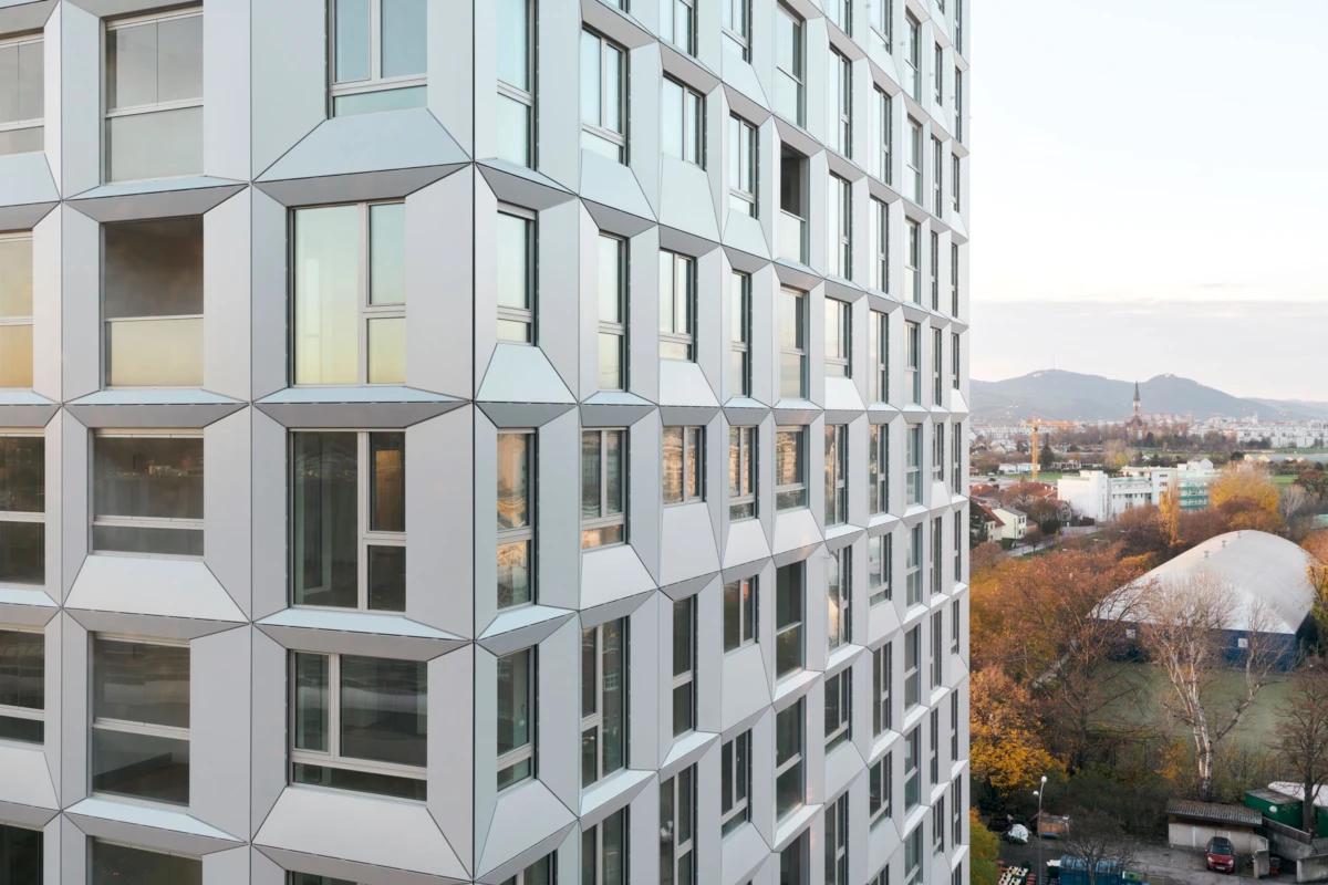 High-rise building exterior with diamond-patterned metal panels, overlooking a cityscape with autumn trees and distant hills.