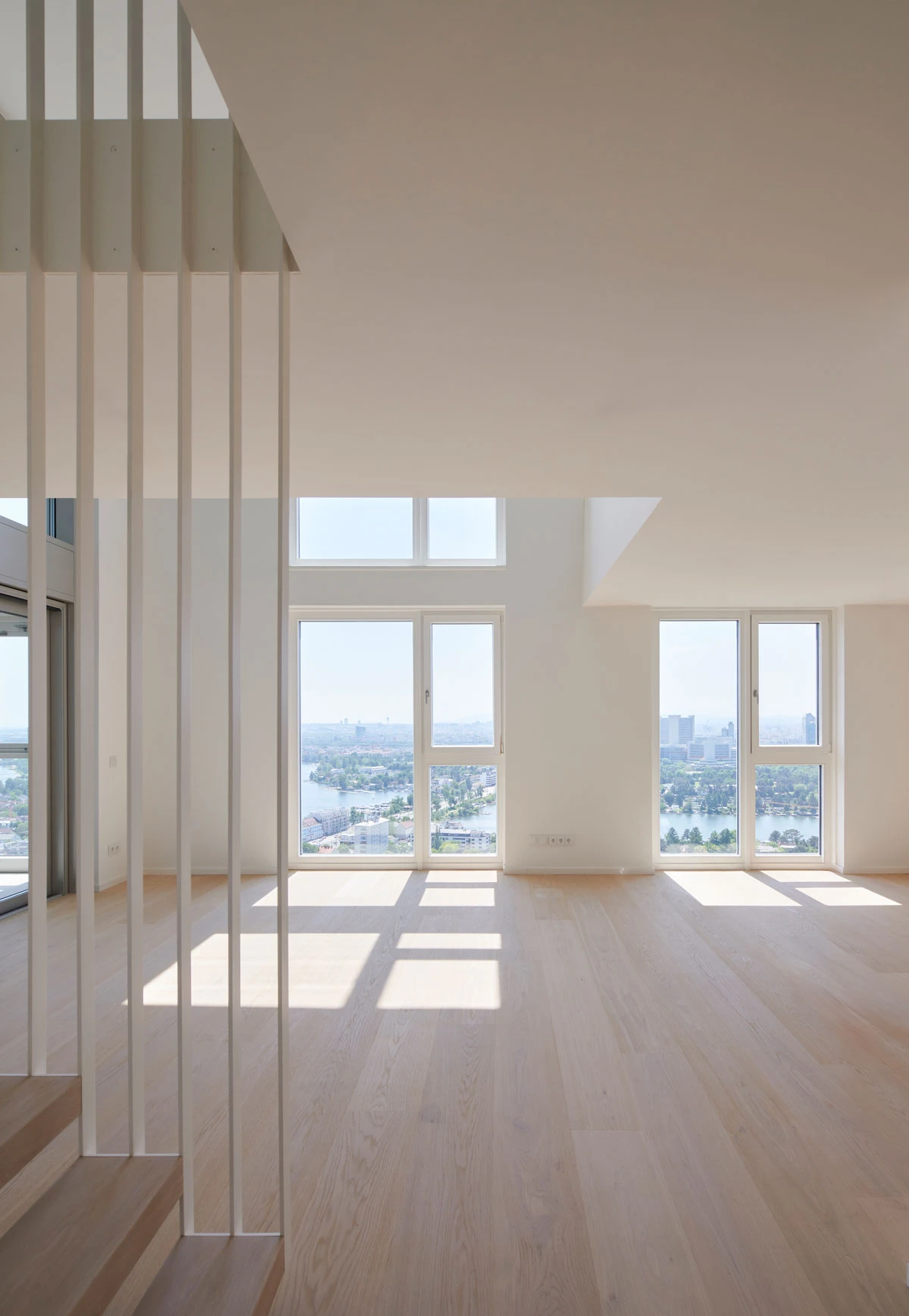 Bright modern open-plan living room with tall windows; sunlight casts rectangular shadows on light wooden floor near a white stair railing.